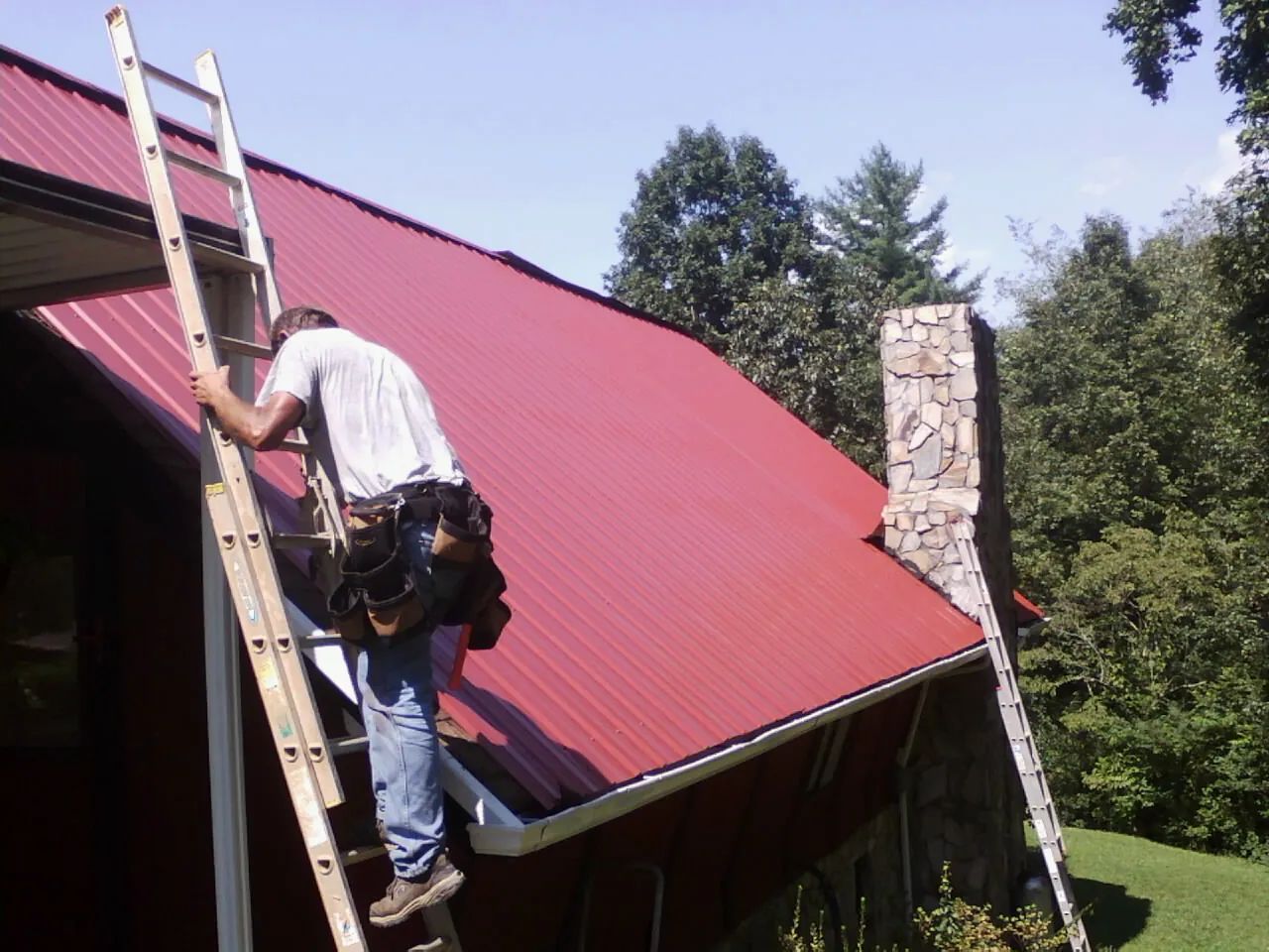 Roofer on a ladder beside a red metal roof and stone chimney — Soffit & Fascia Repair by Roofing Brothers in Howard Beach
